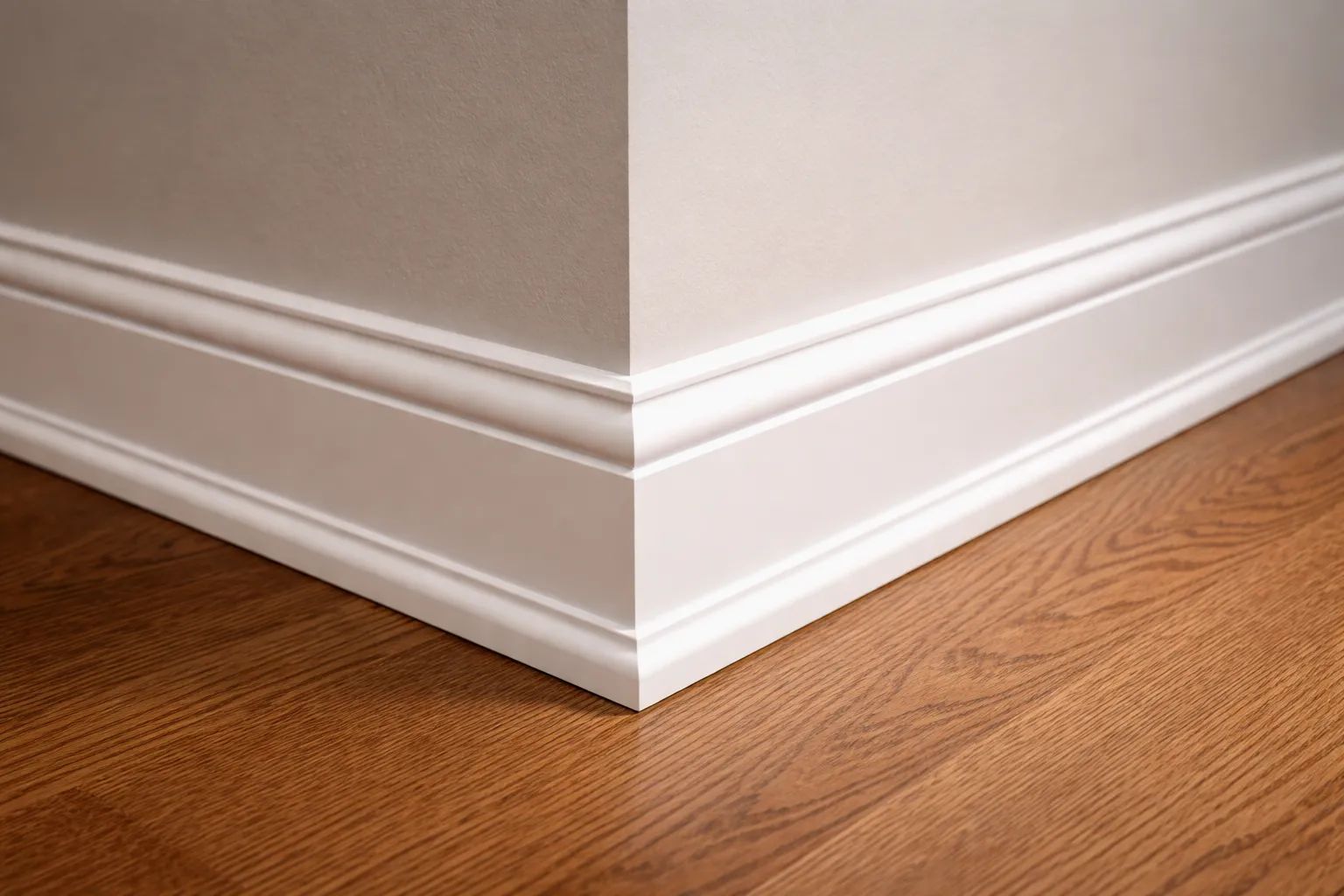 Close up of a clean white baseboard wrapping around an interior wall corner above medium tone hardwood flooring. The smooth joints and crisp profile show precise finish carpentry work.